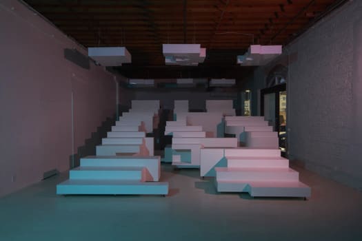 A dim room filled with pale pink and blue stepped seating blocks arranged like bleachers under exposed ceiling ducts.