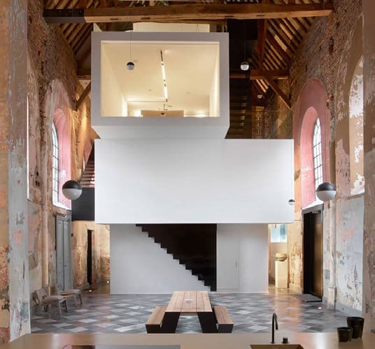 A freestanding white box with a lit interior room inserted inside the stripped brick nave of a former chapel.