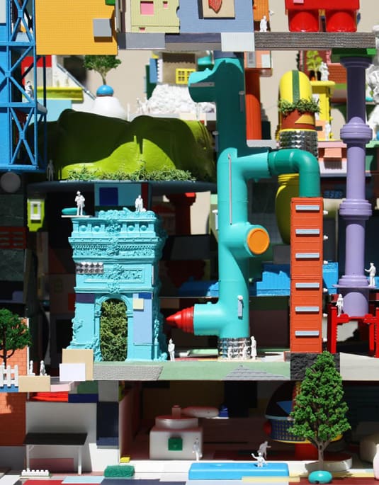 A dense, brightly colored architectural model collaging classical facades, pipes, towers, and miniature figures together.