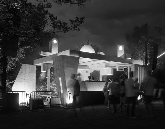 A nighttime outdoor stage built from angled plywood panels and columns, lit from within, with a crowd gathered around it.