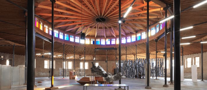 A circular timber-roofed hall ringed with stained glass, columns, and a dark sculptural installation at its center.