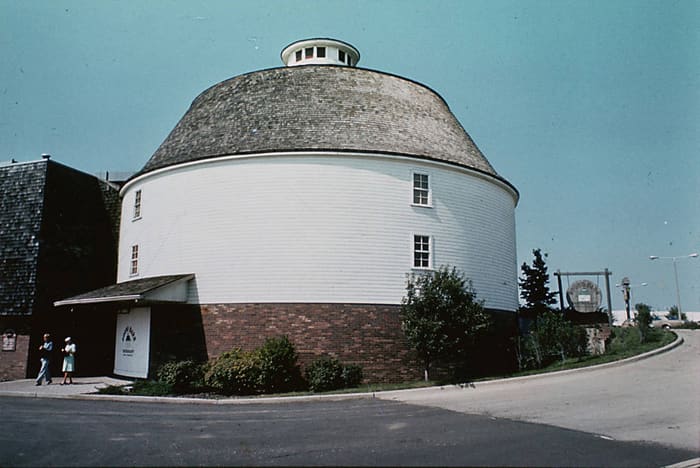A vintage color photo of the white round barn with a shingled dome roof and brick base, two visitors at the entrance.