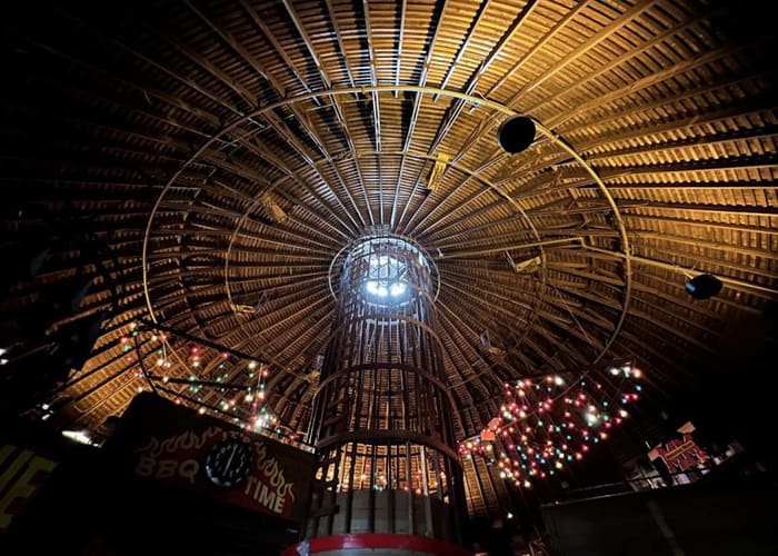 Looking straight up into the round barn's radial timber roof structure, ringed with string lights around a central silo.