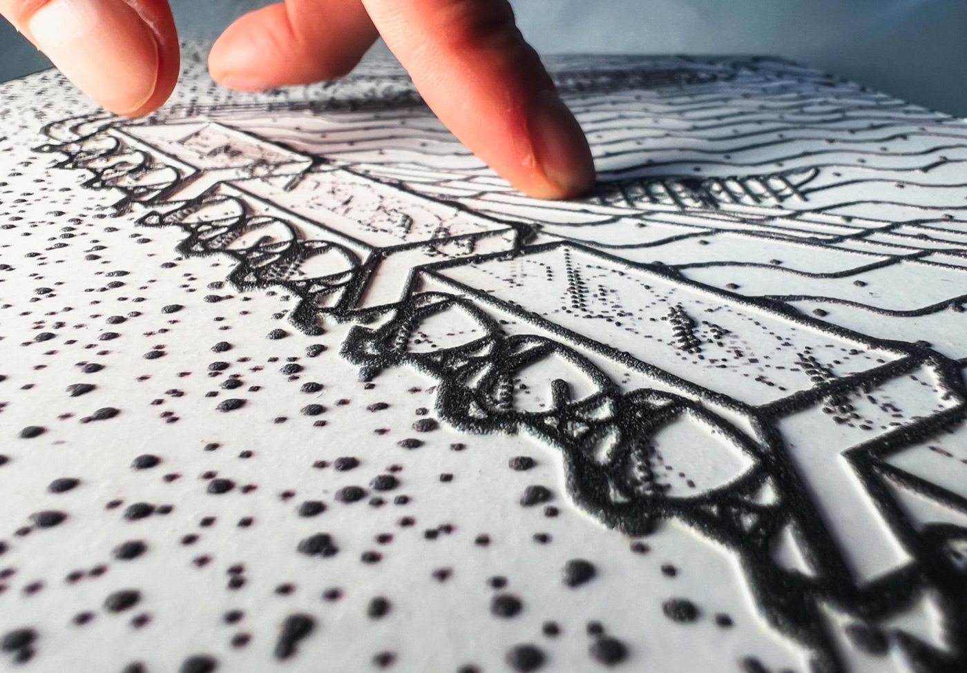Hands reading a swell-paper tactile axonometric drawing; raised black lines form a three-dimensional massing on textured paper amid other tactile sheets.